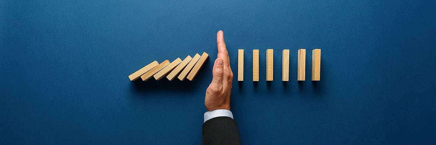 Wide view image of businessman hand stopping collapsing dominos in a conceptual image. Top view over navy blue background.